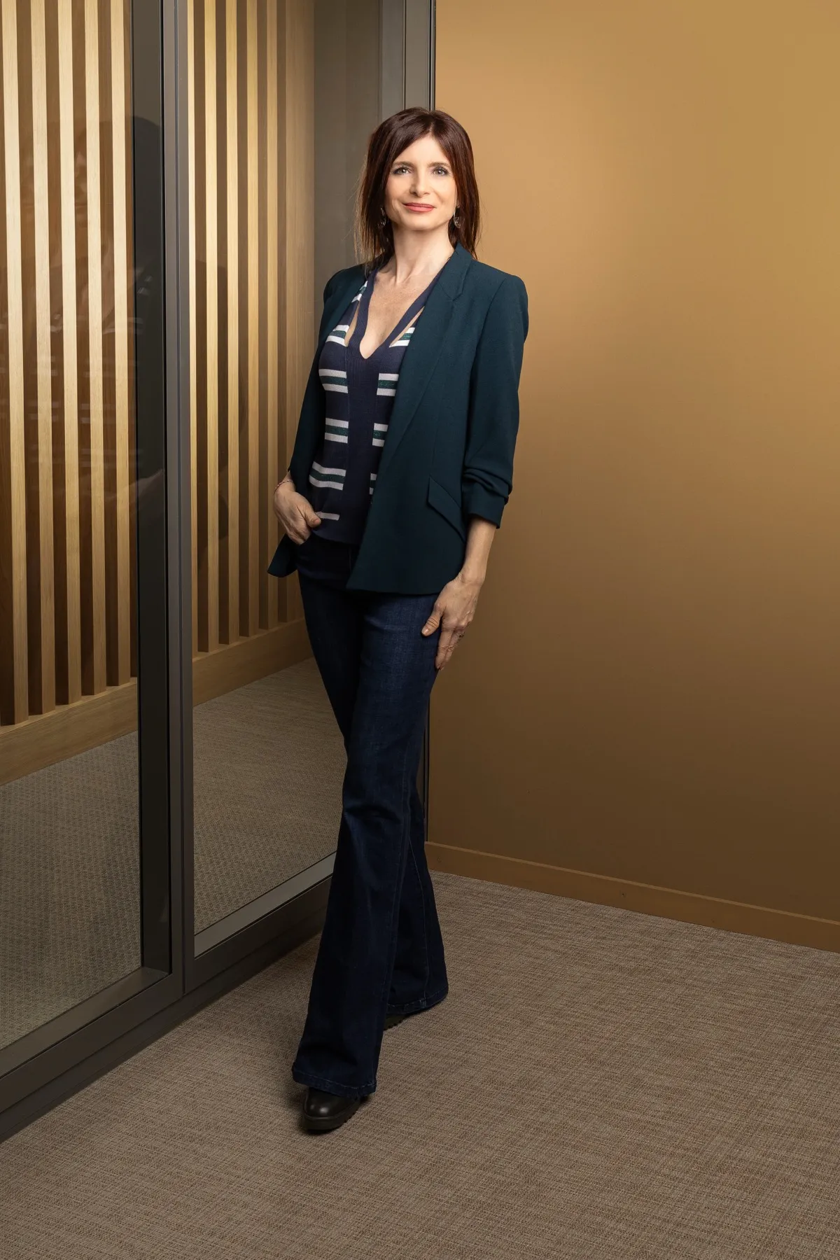 Véronique Pagano, en veste sombre, devant une cloison vitrée aux montants métalliques fins et un mur de lattes de bois clair.